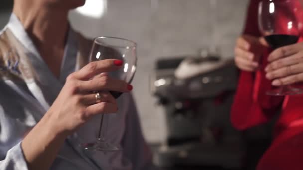 Δύο γυναίκες που κουβεντιάζουν στην κουζίνα και πίνουν κόκκινο κρασί από το γυαλί-ένα φορώντας μπλε πρωινή ρόμπα, το άλλο κόκκινο φόρεμα ρόμπα-γελώντας και χαμογελαστά — Αρχείο Βίντεο