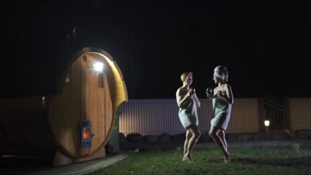 Due ragazze donne che bevono vino rosso da bicchieri all'esterno vicino a una sauna mobile in legno indossando solo asciugamani — Video Stock