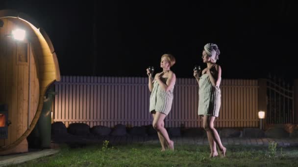 Twee vriendinnen vrouwen drinken rode wijn uit glazen buiten locatie in de buurt van een houten mobiele sauna dragen gewoon handdoeken — Stockvideo