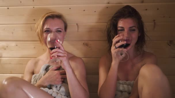 Two beautiful hot sexy girlfriends women drinking red wine from glasses and chatting in wooden finnish Sauna wet from steam - Laughing and smiling — Stock Video
