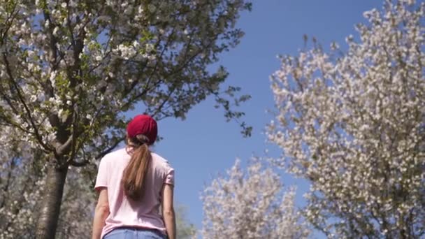 Erfolgreiche Geschäftsfrau genießt ihre Freizeit in einem Park mit blühenden Sakura-Kirschbäumen in Jeans, rosa T-Shirt und modisch roter Mütze — Stockvideo