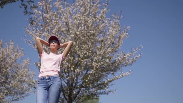 Mulher de negócios bem sucedida desfruta de seu tempo livre de lazer em um parque com árvores florescentes sakura cereja vestindo jeans, camiseta rosa e um boné vermelho moda — Vídeo de Stock