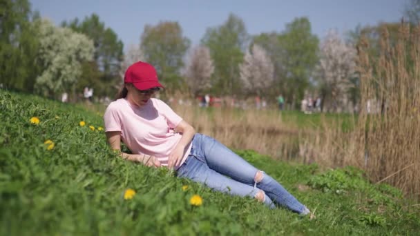 成功的商务女性享受她的休闲空闲时间在公园与开花樱花樱花树穿着牛仔裤，粉红色T恤和时尚红色帽子 — 图库视频影像
