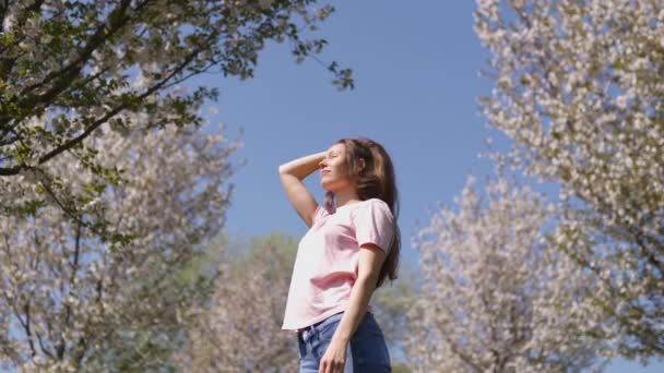 Erfolgreiche Geschäftsfrau genießt ihre Freizeit in einem Park mit blühenden Sakura-Kirschbäumen in Jeans, rosa T-Shirt und modisch roter Mütze — Stockvideo