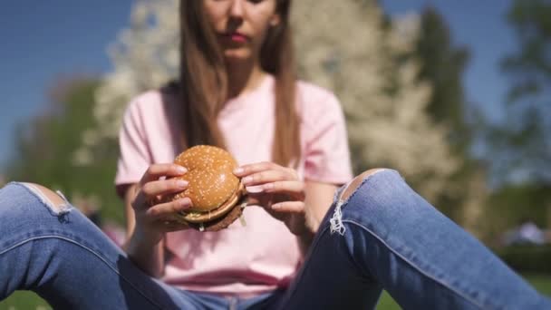 Erfolgreiche Geschäftsfrau, die Fast-Food-Burger Cheesburger isst, genießt ihre Freizeit in einem Park mit blühenden Sakura-Kirschbäumen in Jeans, rosa T-Shirt und modisch roter Mütze — Stockvideo