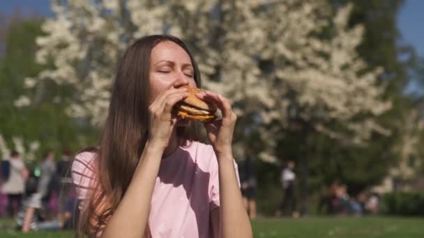 ファーストフードバーガーチーズバーガーを食べる成功したビジネスウーマンは、ジーンズ、ピンクのTシャツとファッションの赤い帽子を着て桜の木を咲かせる公園で彼女の余暇の時間を楽しんでいます — ストック動画
