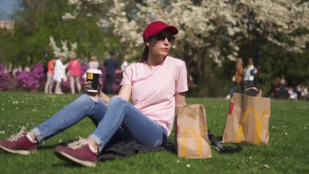 Ρίγα, Λετονία-28 Απριλίου, 2019: επιτυχημένη επιχείρηση γυναίκα που τρώει ΜακΝτόναλντς Big Mac Burger τσίζμπεργκερ και πίνοντας Κόκα κόλα απολαμβάνει τον ελεύθερο χρόνο της σε ένα πάρκο — Αρχείο Βίντεο