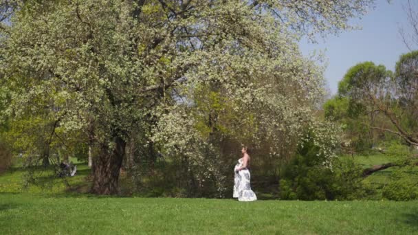 Junge Reisende schwangere Frau geht, rennt, dreht sich um und genießt ihre Freizeit in einem Park mit blühenden Sakura-Kirschbäumen in einem sommerlichen hellen langen Kleid mit Blumenmuster — Stockvideo