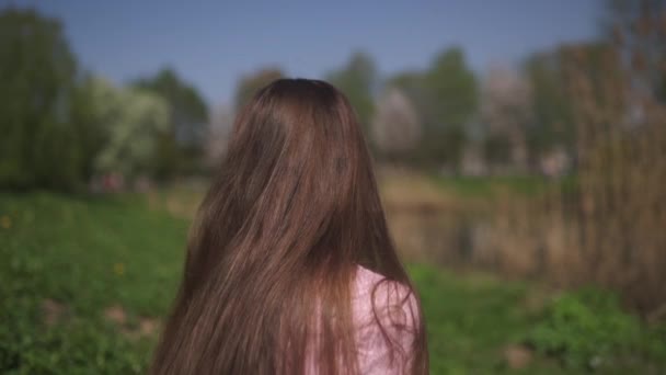 MOCIÓN LENTA 120fps: Joven viajero feliz morena niña de pelo castaño sonriendo y dando la vuelta en un nuevo país de destino con un sakura rosa árboles de flor de cerezo en los estados bálticos - Cabello volador — Vídeos de Stock