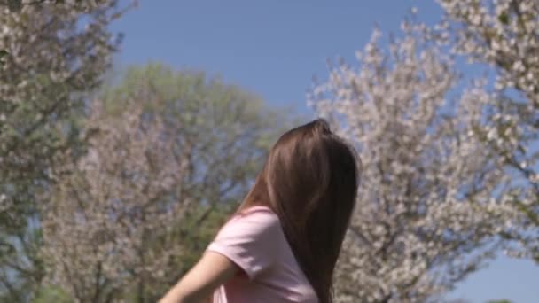 Zeitlupe 120fps: junge, glückliche Frau mit braunhaarigen Haaren lächelt und dreht sich um in einem neuen Zielland mit rosa Sakura-Kirschblütenbäumen in den baltischen Staaten - fliegendes Haar — Stockvideo