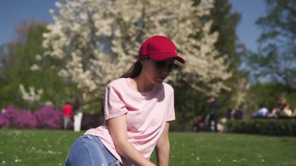 成功的商务女性享受她的休闲空闲时间在公园与开花樱花樱花树穿着牛仔裤，粉红色T恤和时尚红色帽子 — 图库视频影像
