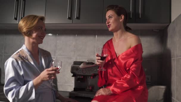 Duas mulheres conversando na cozinha e beber vinho tinto de vidro - Um vestindo vestido de manhã azul, o outro vestido de roupão vermelho - rindo e sorrindo — Vídeo de Stock