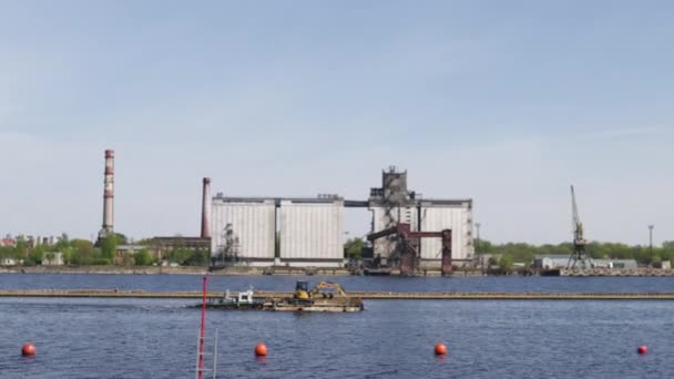 Říční člun se rozjede podél řeky Daugava s escavátorem na palubě-slunečné počasí s přístavem na pozadí — Stock video
