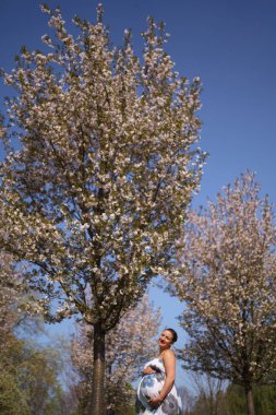 Mutlu genç yakında anne olmak - Genç gezgin hamile kadın çiçek açan sakura kiraz ağaçları çiçek desenli bir yaz ışık uzun elbise giyen bir parkta boş zaman sahiptir