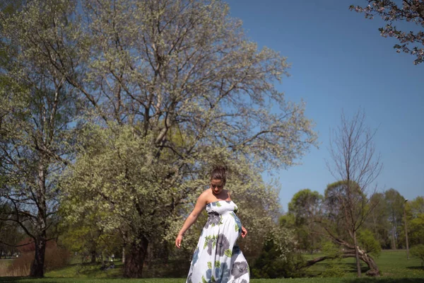 Jeune voyageuse enceinte marchant, courant, se retournant et jouissant de son temps libre dans un parc avec des cerisiers sakura en fleurs portant une robe longue lumière d'été avec motif de fleurs — Photo