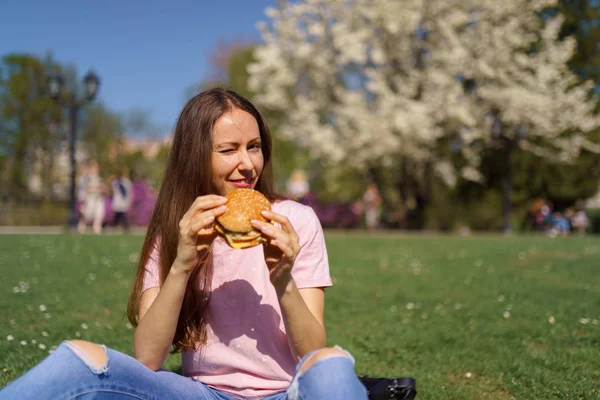 Успішна бізнес-леді, яка їсть фастфуд бургер, насолоджується вільним дозвіллям у парку з квітучими сакура вишневими деревами в джинсах, рожевій футболці та модній червоній шапці — стокове фото