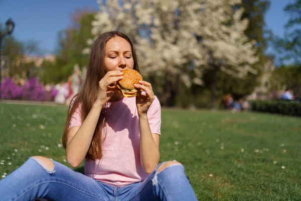 Успішна бізнес-леді, яка їсть фастфуд бургер, насолоджується вільним дозвіллям у парку з квітучими сакура вишневими деревами в джинсах, рожевій футболці та модній червоній шапці — стокове фото
