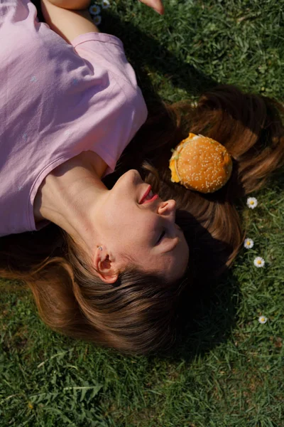 Успішна бізнес-леді лежить на траві з фаст-фудом бургер чізбургер на волоссі насолоджуючись дозвіллям вільний час у парку з квітучими сакура вишневими деревами в рожевій футболці — стокове фото