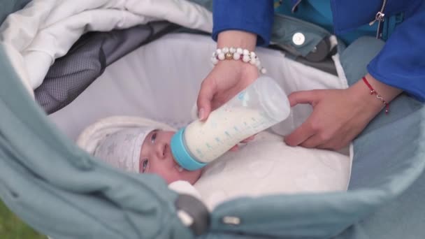 Moeder voedt baby jongen peuter kind melk uit een plastic fles met een helder blauwe tepel-Kaukasische Wittekind met een ouders hand zichtbaar-leggen in een baby vervoer met witte lakens — Stockvideo