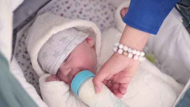어머니는 밝은 파란색 젖꼭지가있는 플라스틱 병에서 아기 소년 유아 용 우유를 공급합니다 - 부모의 손이 보이는 백인 흰색 어린이 - 흰색 시트가있는 유모차에 누워 — 비디오