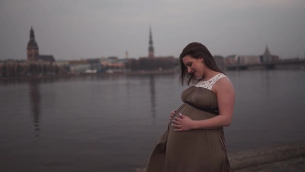 Držet si břicho zblízka-Mladá těhotná žena je šťastná v zemi svého cestovního cíle Lotyšsko s výhledem na město Riga a řeka Daugava-bílá Kavkazská dívka s mateřskými šaty — Stock video