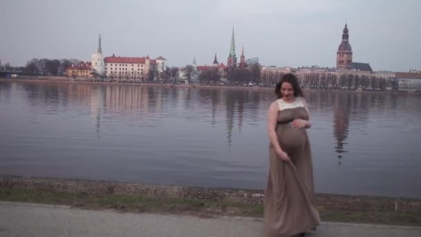 Feliz baile - Mujer embarazada joven es feliz en su país de destino Letonia con una vista sobre la ciudad de Riga y el río Daugava - Chica blanca caucásica con vestido de maternidad — Vídeo de stock