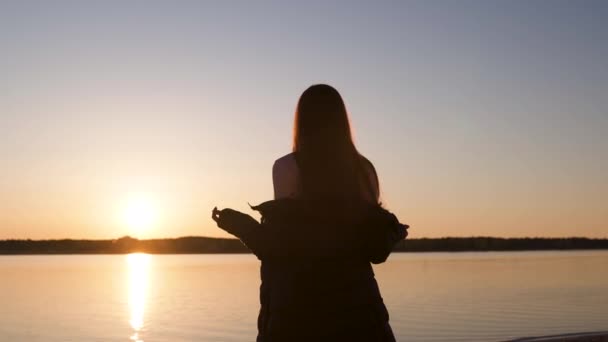 Ung kvinna njuter av solnedgången under Golden Hour på en flod strand på våren bär vita byxor, svettkort och jacka - kaukasisk vit flicka är en glad resenär — Stockvideo