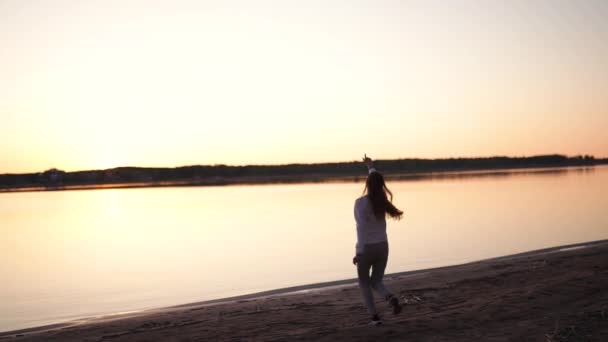 Lacht lustig und zeigt auf eine untergehende Sonne - junge Frau genießt Sonnenuntergang zur goldenen Stunde am Flussstrand im Frühling in weißer Hose, Jogginghose und Jacke - kaukasisches weißes Mädchen — Stockvideo