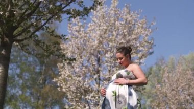 Mutlu genç yakında anne olmak - Genç gezgin hamile kadın çiçek açan sakura kiraz ağaçları çiçek desenli bir yaz ışık uzun elbise giyen bir parkta boş zaman sahiptir