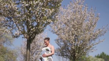 Mutlu genç yakında anne olmak - Genç gezgin hamile kadın çiçek açan sakura kiraz ağaçları çiçek desenli bir yaz ışık uzun elbise giyen bir parkta boş zaman sahiptir