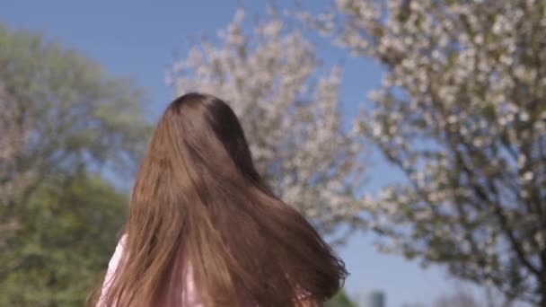 MOCIÓN LENTA 120fps: Joven viajero feliz morena niña de pelo castaño sonriendo y dando la vuelta en un nuevo país de destino con un sakura rosa árboles de flor de cerezo en los estados bálticos - Cabello volador — Vídeo de stock