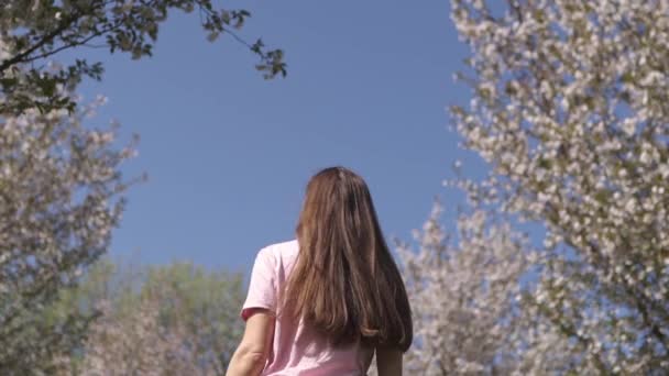 MOCIÓN LENTA 120fps: Joven viajero feliz morena niña de pelo castaño sonriendo y dando la vuelta en un nuevo país de destino con un sakura rosa árboles de flor de cerezo en los estados bálticos - Cabello volador — Vídeo de stock