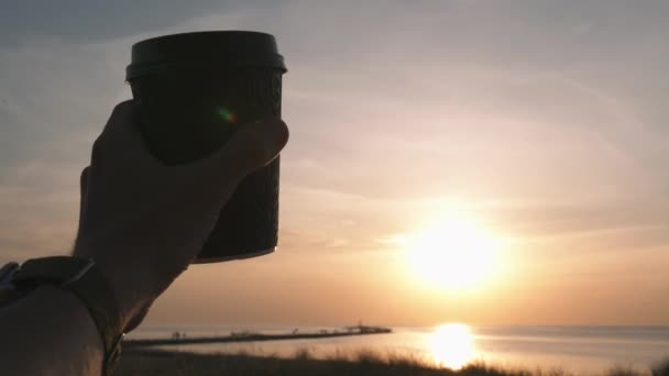 Gelukkige reiziger man hand houden zwart papier kopje koffie wijst naar de zonsondergang en het instellen van zon op het strand — Stockvideo