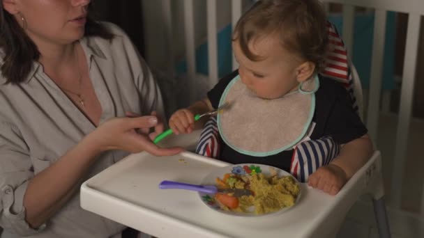 Μητέρα σίτιση χυλό για το μωρό παιδί της εσωτερικό παιδικό δωμάτιο-νεαρή μαμά σε γκρίζο σπίτι φορούν και ο γιος της τρώγοντας κάθονται στο κάθισμα του μωρού-χαρούμενοι χαμογελαστοί άνθρωποι — Αρχείο Βίντεο