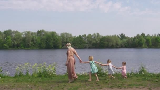 Junge blonde Hippie-Mutter, die Zeit mit ihren kleinen Mädchen in einem Park hat - Töchter tragen ähnliche Kleider mit Erdbeerprint - laufen wie Gänse in einer Reihe am Fluss entlang — Stockvideo