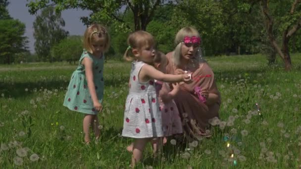 Jovem loira hippie mãe ter tempo de qualidade com suas meninas em um parque soprando bolhas de sabão - Filhas usam vestidos semelhantes com impressão de morango - Conceito de valores familiares — Vídeo de Stock