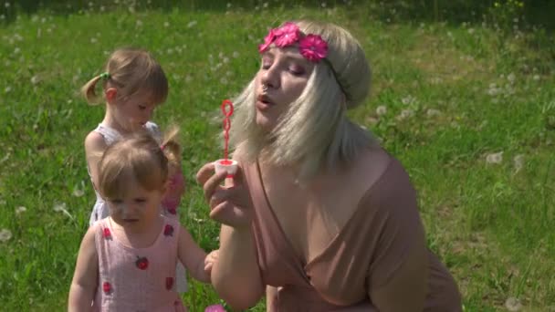 Ung blond hippie mamma har kvalitetstid med sina små flickor i en park blåser såpbubblor - Döttrar bär liknande klänningar med jordgubbstryck - Familjevärderingar koncept — Stockvideo