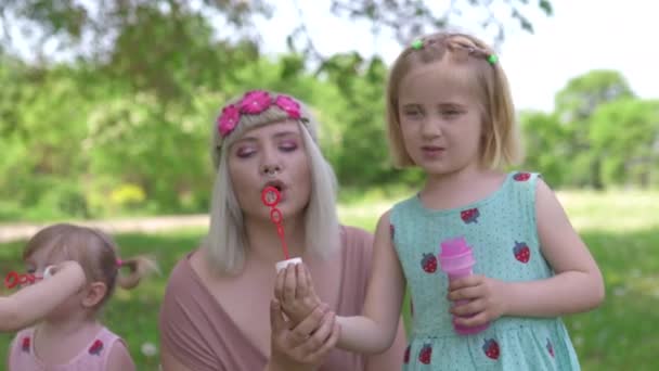 Молода блондинка хіпі мати проводить якісний час зі своїми дівчатами в парку дме мильні бульбашки Дочки носять схожі сукні з полуничним принтом концепція сімейних цінностей — стокове відео