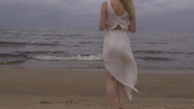 Behind view - Beautiful young blonde woman walking toward water beach nymph in white dress near sea with waves during a dull gloomy weather with stormy wind and rain