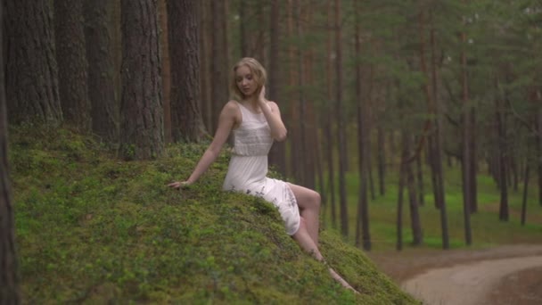 Gyönyörű fiatal szőke nő ül az erdőben nimfa fehér ruhában az örökzöld fa