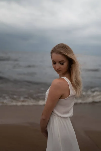 Primer plano retrato de hermosa joven rubia playa ninfa en vestido blanco cerca del mar con olas durante un clima sombrío aburrido con viento tormentoso y lluvia — Foto de Stock