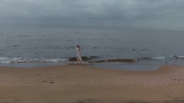 Havadan yan görünüm - Fırtınalı rüzgar ve yağmur ile sıkıcı kasvetli havalarda dalgalar ile deniz kenarında deniz kenarında beyaz elbise li bir plaj perisi üzerinde deniz boyunca yürüyen güzel genç sarışın kadın