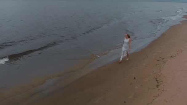 Luftaufnahme - schöne junge blonde Frau, die am Meer entlang geht, an einem Strand Nymphe in weißem Kleid in der Nähe des Meeres mit Wellen während eines trüben Wetters mit stürmischem Wind und Regen — Stockvideo