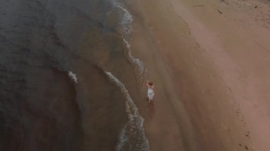 Havadan üst görünümü - Fırtınalı rüzgar ve yağmur ile donuk kasvetli havalarda dalgalar ile deniz kenarında beyaz elbiseli bir plaj perisi üzerinde deniz boyunca yürüyen güzel genç sarışın kadın