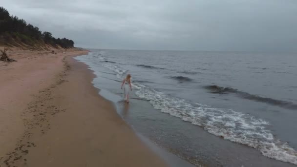 Аерофотозйомка - Красива молода блондинка, що йде уздовж моря на пляжі німфа в білій сукні біля моря з хвилями під час нудної похмурої похмурої погоди з штормовим вітром і дощем — стокове відео