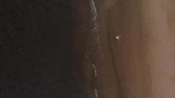 Vista aérea superior - Linda jovem loira andando ao longo do mar em uma ninfa de praia em vestido branco perto do mar com ondas durante um clima sombrio maçante com vento tempestuoso e chuva — Vídeo de Stock