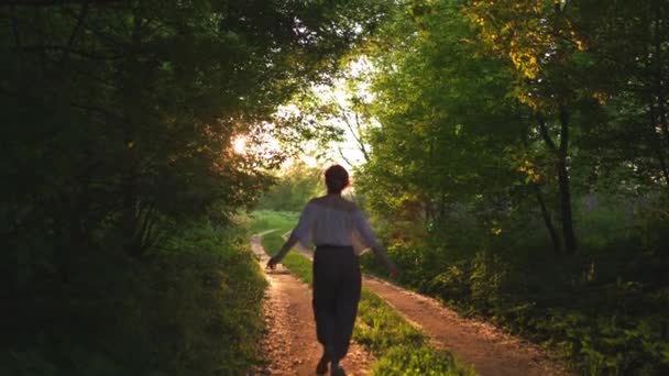 Redhead kvinna i byxor och vit skjorta Walking-Sunset land Off Road med vackra kvällssol ljusstrålar, gröna löv träd runt-naturen är ett bra ställe att koppla av i bakgrunden — Stockvideo