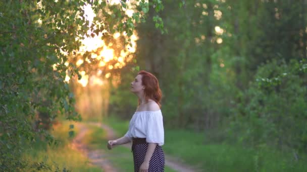 Mujer pelirroja en pantalones y camisa blanca posando - Campo al atardecer fuera de la carretera con hermosos rayos de luz del sol por la noche, árboles de hojas verdes alrededor - La naturaleza es un gran lugar para relajarse en el fondo — Vídeos de Stock