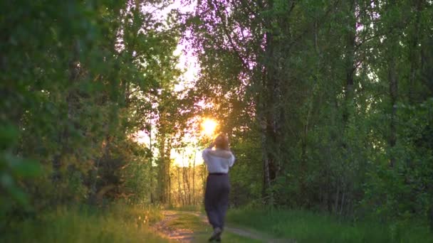 Redhead kvinna i byxor och vit skjorta Walking-Sunset land Off Road med vackra kvällssol ljusstrålar, gröna löv träd runt-naturen är ett bra ställe att koppla av i bakgrunden — Stockvideo
