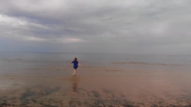 Aerial: Giovane donna in abito blu brillante in esecuzione - acqua bassa scenico paesaggio giallastro tramonto al Mar Baltico - Drone vista volante dall'alto — Video Stock